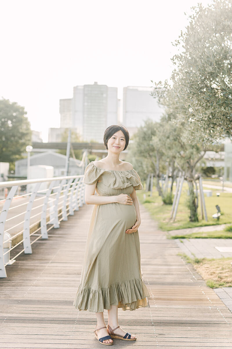 Woman placing her hands on her belly and looking at the camera, wearing her green khaki dress in a light and airy lighting. Beautiful maternity photograph.