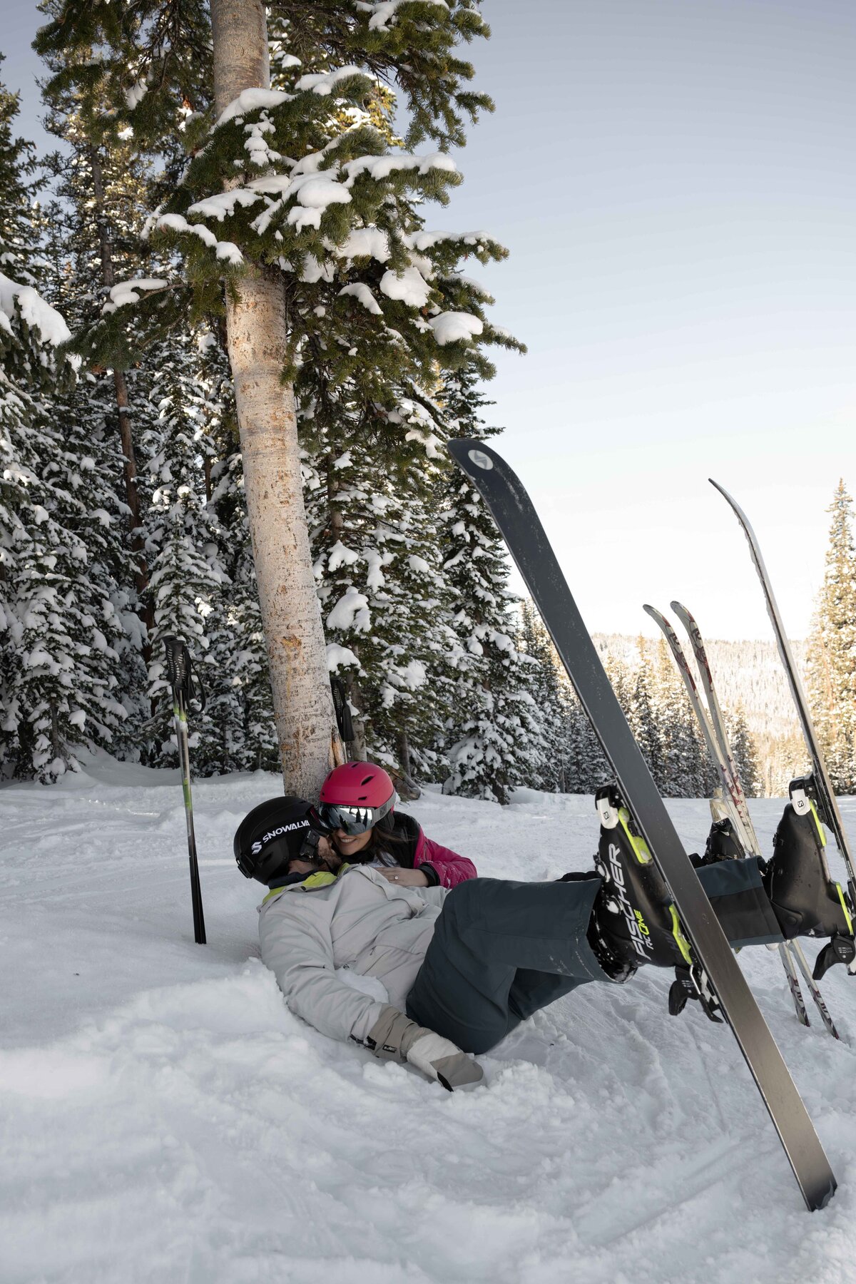 "Playful and romantic moment of a couple lying in the snow after skiing in Vail, Colorado, surrounded by snow-covered trees and breathtaking mountain scenery. This adventure elopement captures the fun, intimacy, and spontaneity of a winter wedding experience in the Rocky Mountains.