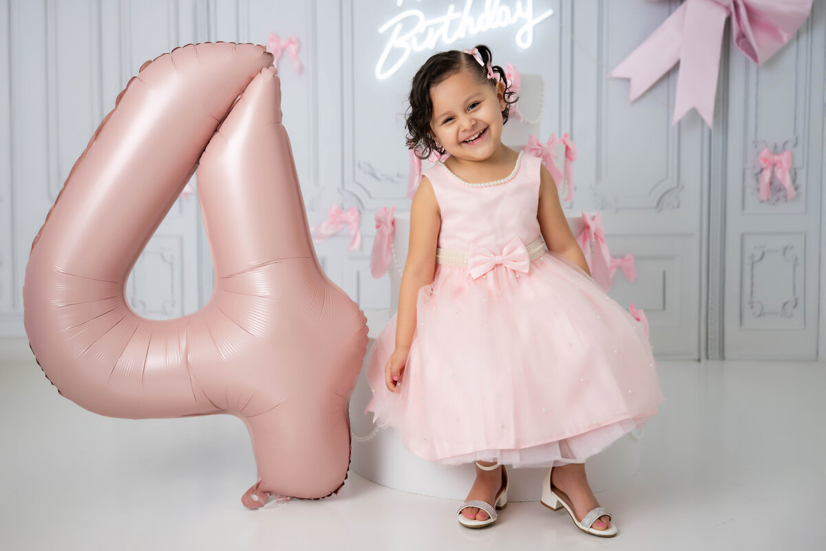 Toddler sitting next to a giant pink number four with a neon sign and bows in the background. 