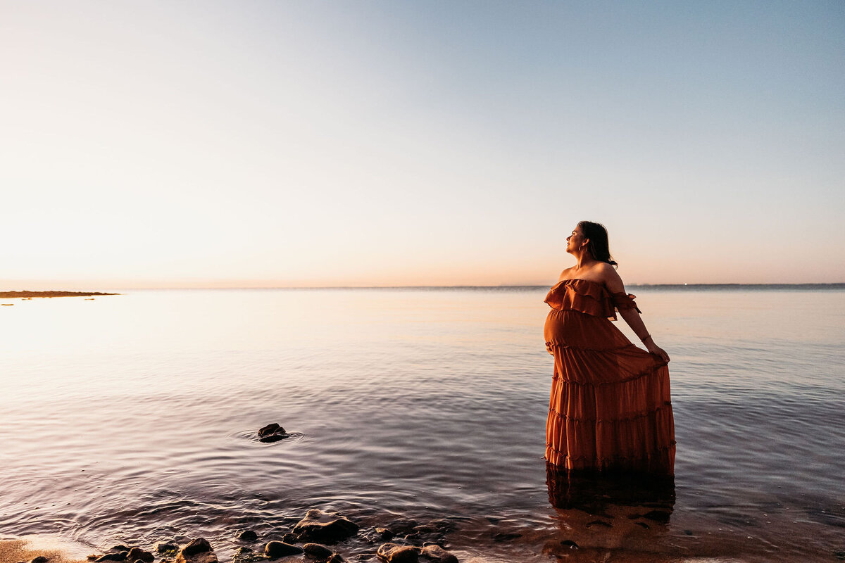 Pregnant woman wearing red gown stands in water at maternity photography session