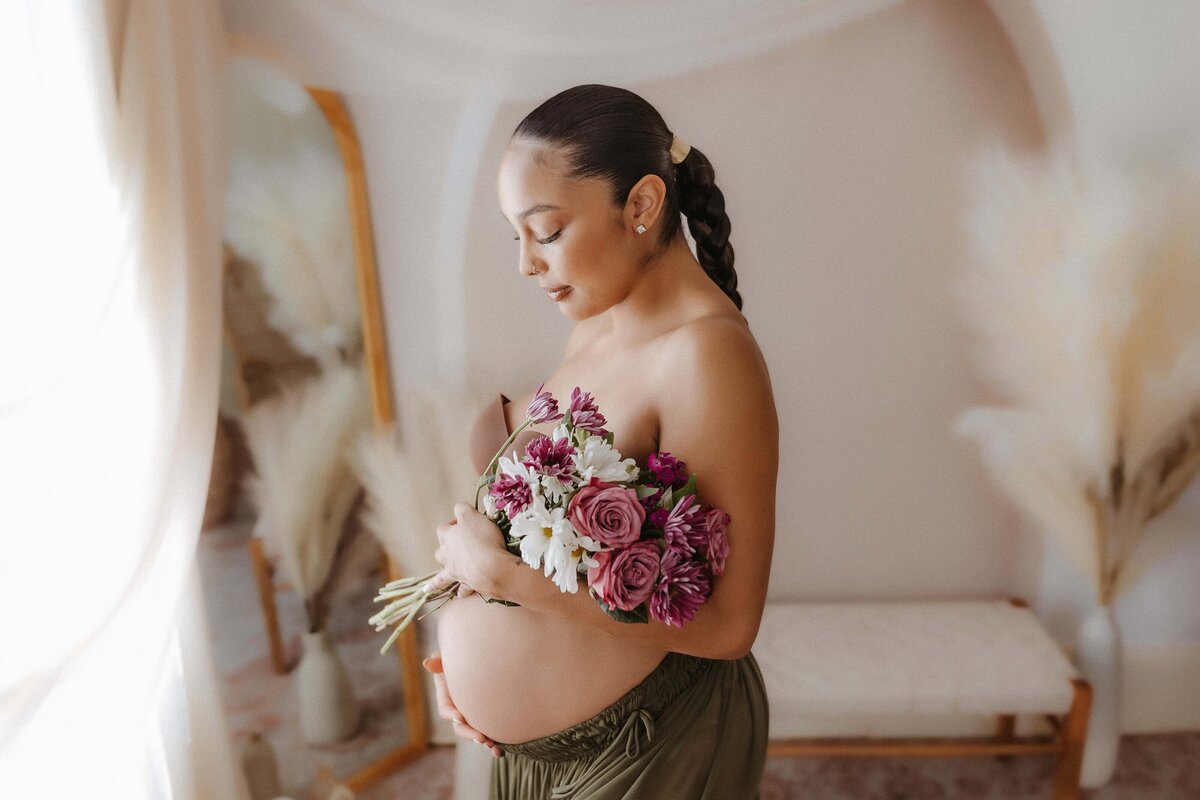 Pregnant woman in a dimly lit room holding a bouquet of purple and white flowers, standing sideways, with curtains and a mirror in the background.