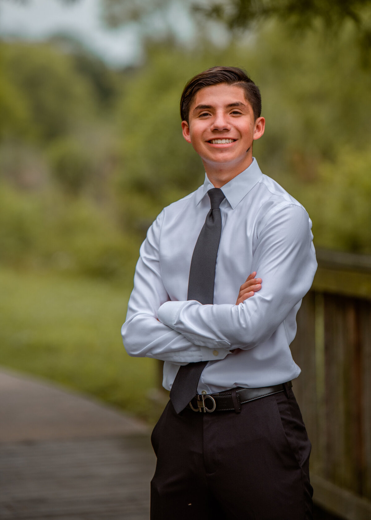 A Pearland senior stands on a wooden bridge with his arms folded while he smiles at the camera.