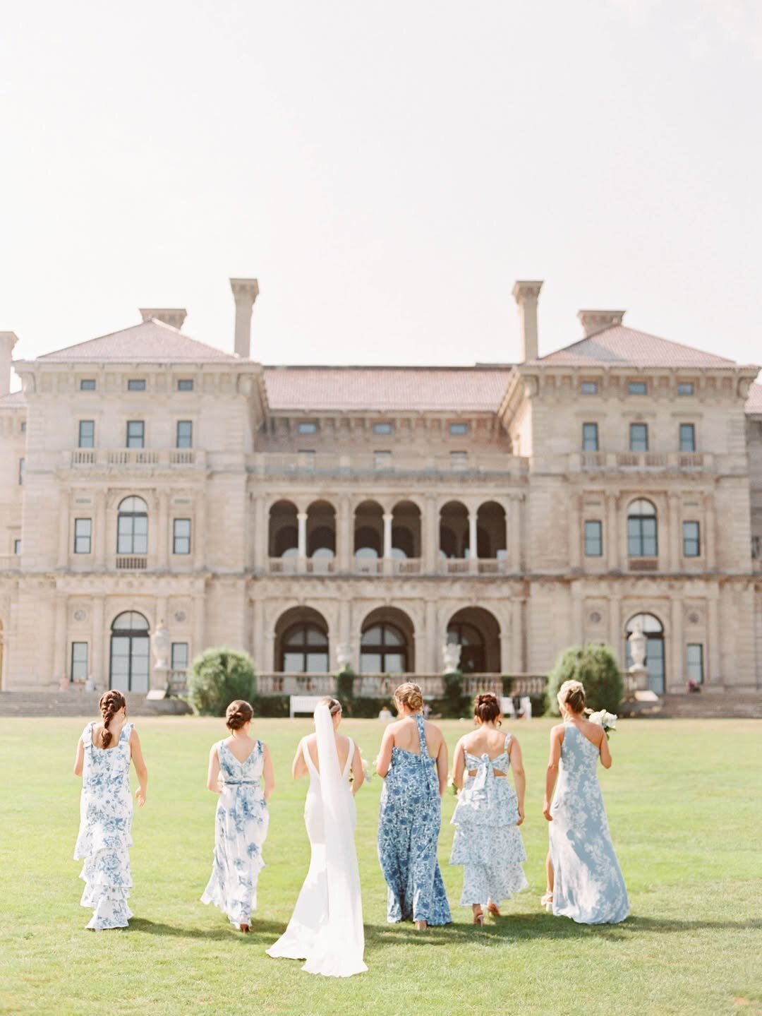Wedding Photographer RI | Bridesmaids in blue floral dresses and a bride in white walk towards a large, elegant mansion with arches. 