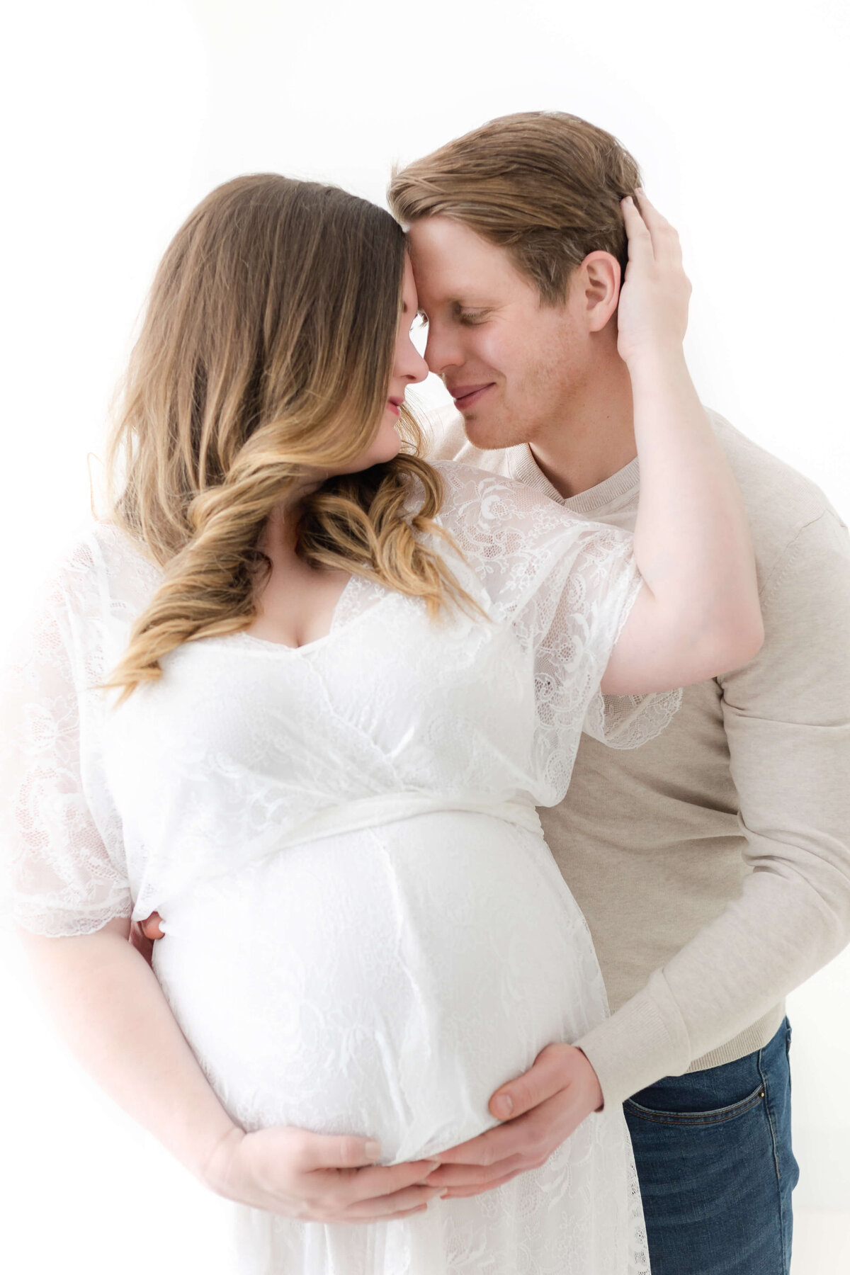 Pregnant mama in white lace dress in studio with her husband holding a hand on her belly. Captured by Collingwood Maternity Photographer Jennifer Walton