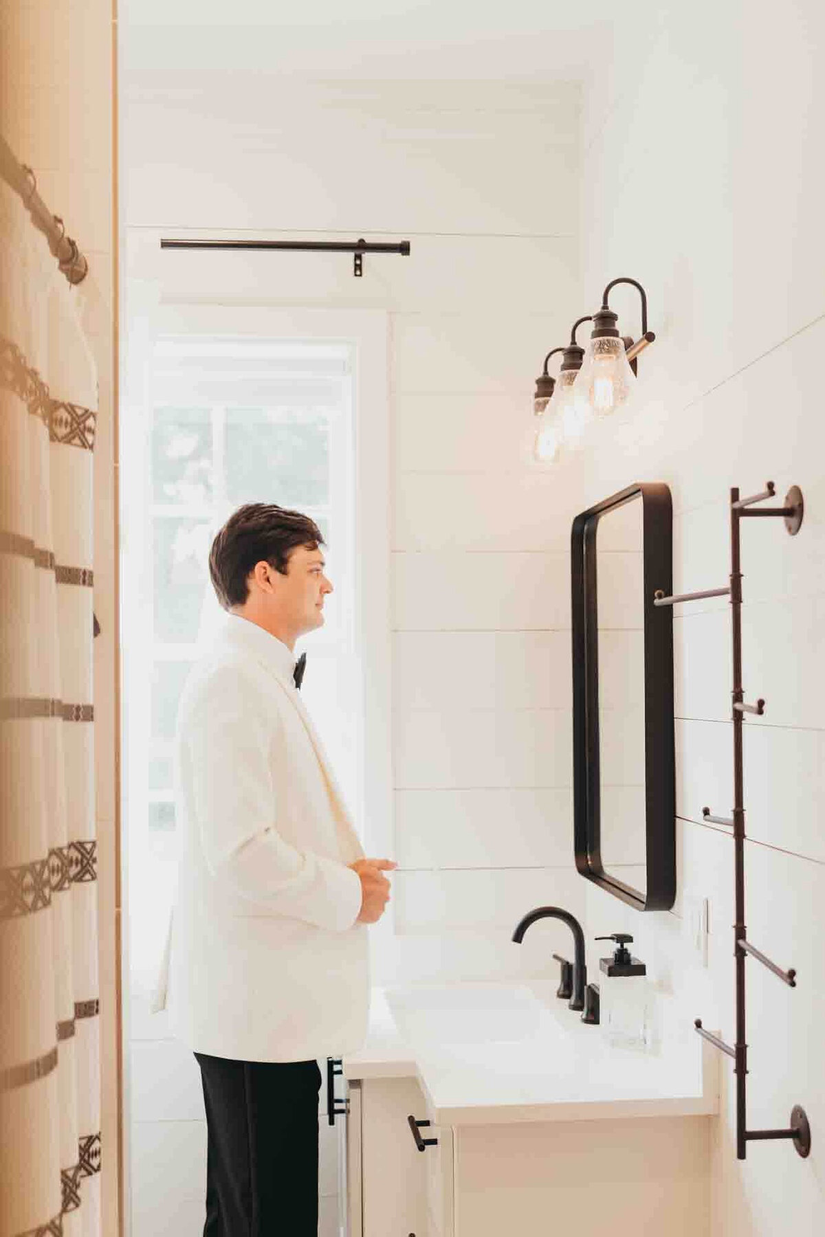 groom looks into black frame mirror in white bathroom, fixing his white suit before his wedding.