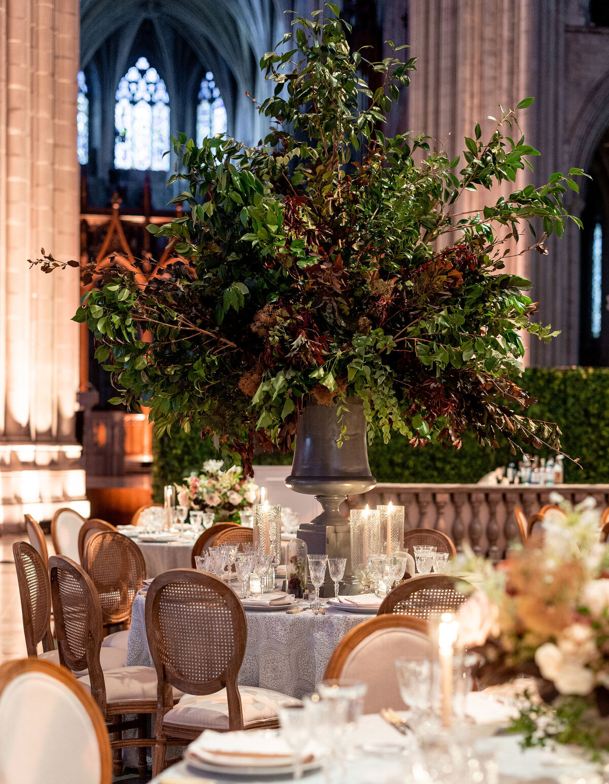 An elegantly set table is displayed in a grand, cathedral-like hall at Washington National Cathedral in Washington D.C., captured by photographer Twah Dougherty. The table features a large floral arrangement with lush greenery and branches in a tall vase. Tall candles, fine glassware, and upholstered wooden chairs adorn the setting, all under the soft glow of ambient lighting.