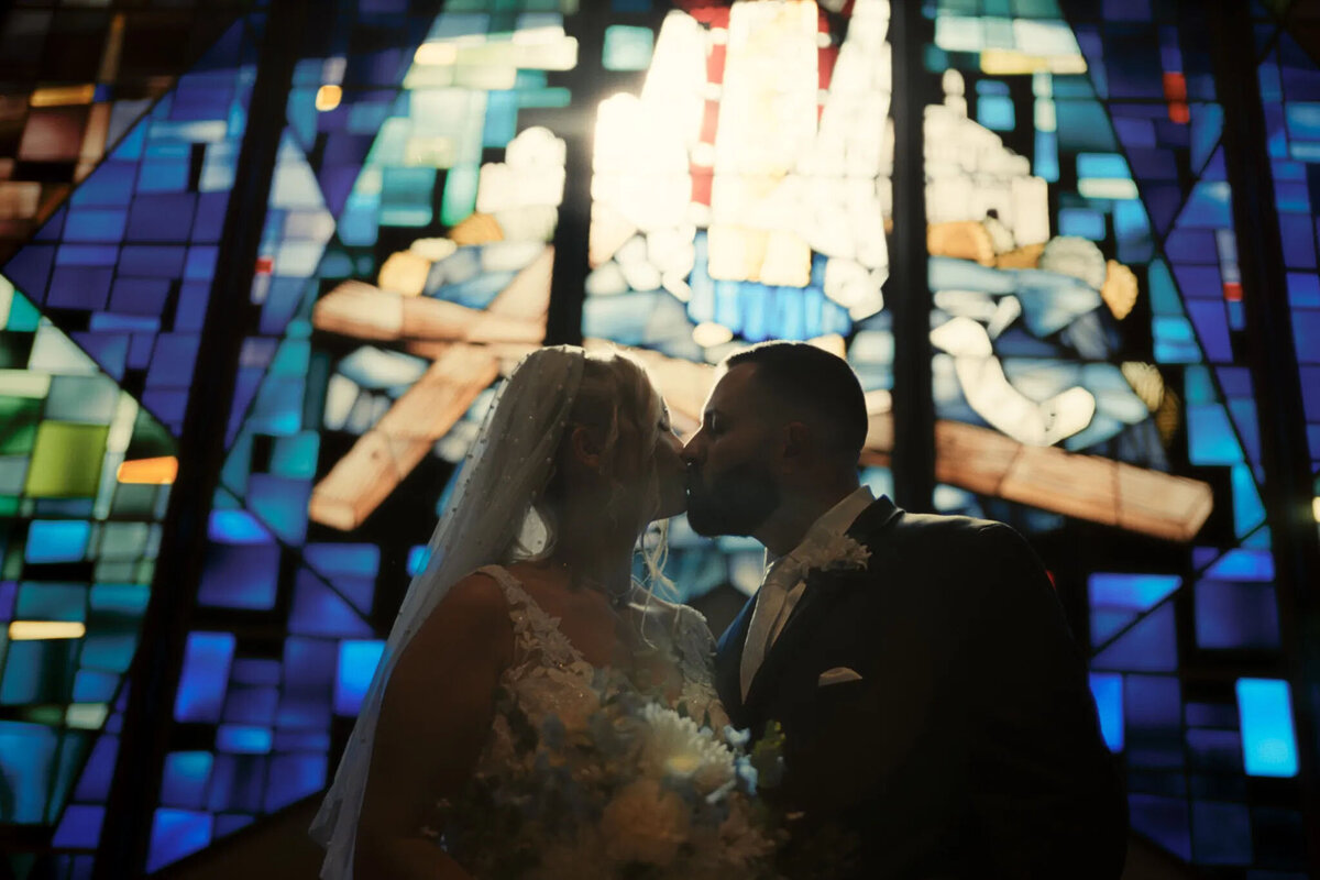 A bride and groom share a kiss in front of a vibrant stained glass window inside a church, with soft blue and purple light illuminating the scene—captured beautifully by an experienced NJ wedding photographer.
