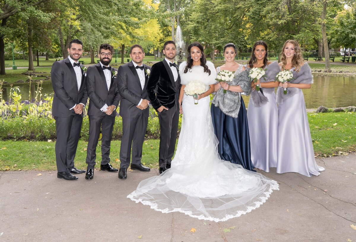 The bridal party is posing together in a lush park setting, showcasing a joyful group shot. The bride and groom stand at the center with their bridesmaids and groomsmen around them, all dressed in their wedding attire. The picturesque park backdrop complements their elegant outfits and the overall celebratory mood of the occasion.