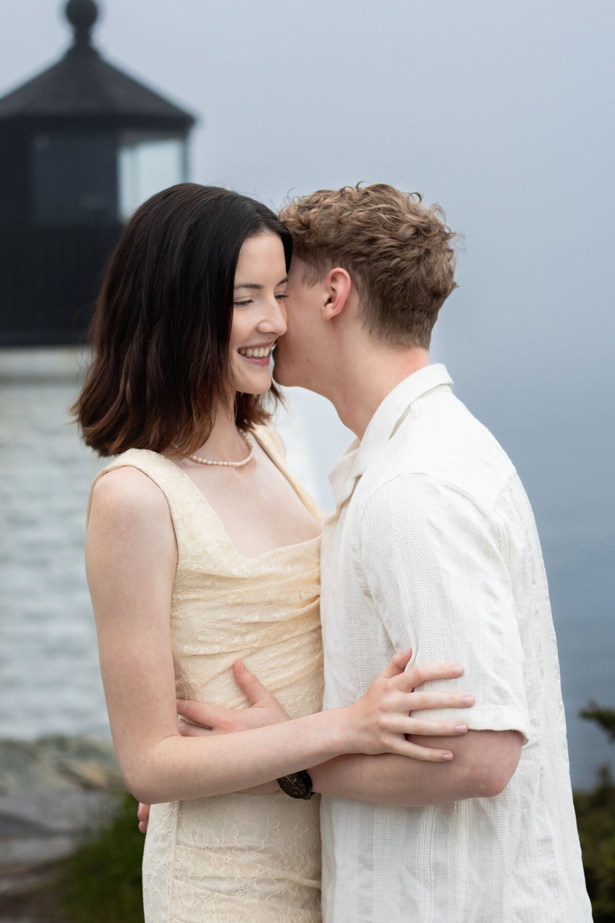 Wedding Photographers in RI | Kelsey Sheehan Photography Timeless Rhode Island Weddings | A couple embracing near an outdoor wall. The woman is smiling, wearing a light dress; the man leans in closely, wearing a white shirt. The mood is affectionate.