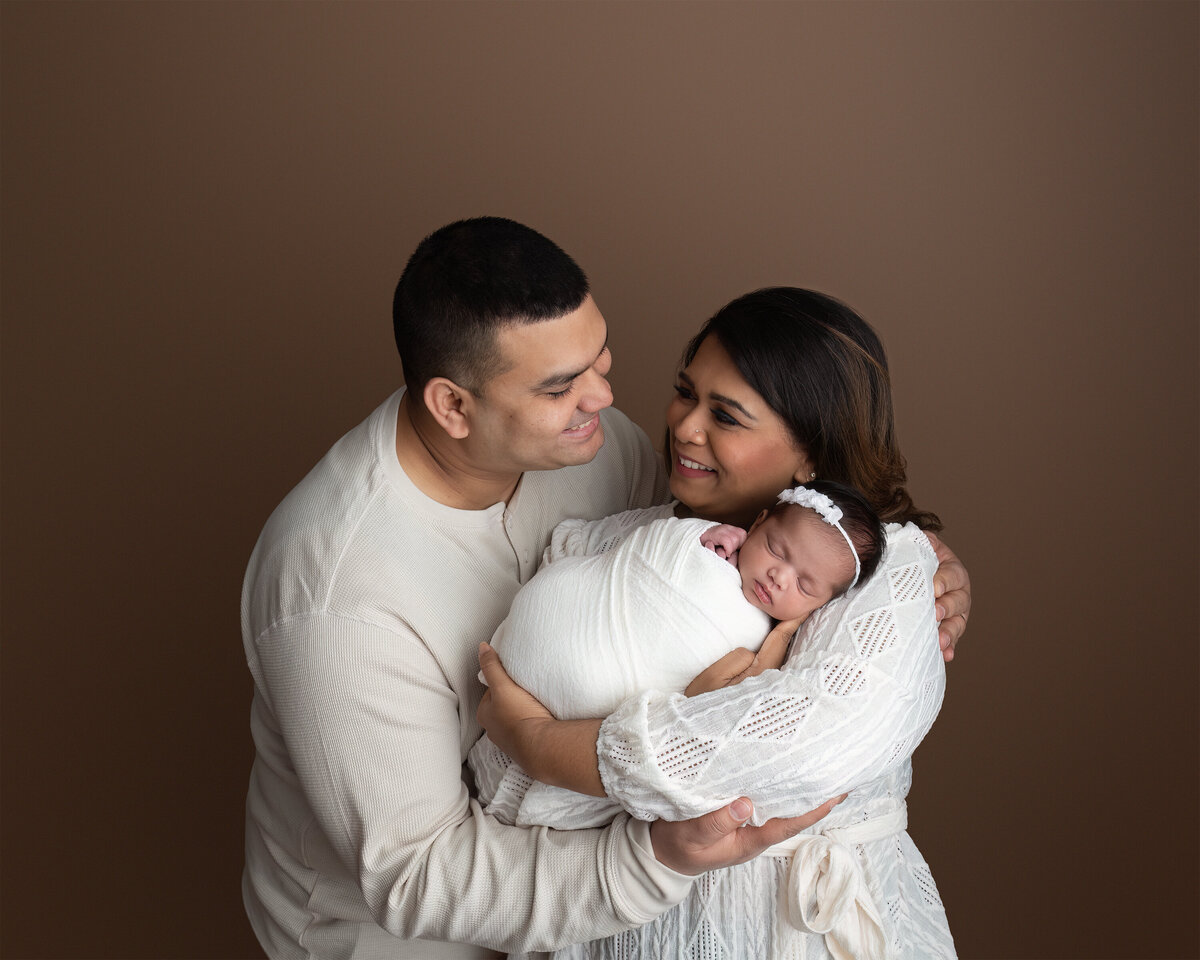 A warm and intimate family portrait captured in-studio, featuring both parents holding their newborn daughter swaddled in white. Photographed at Meraki Photographic Creations in Paris, Ontario.