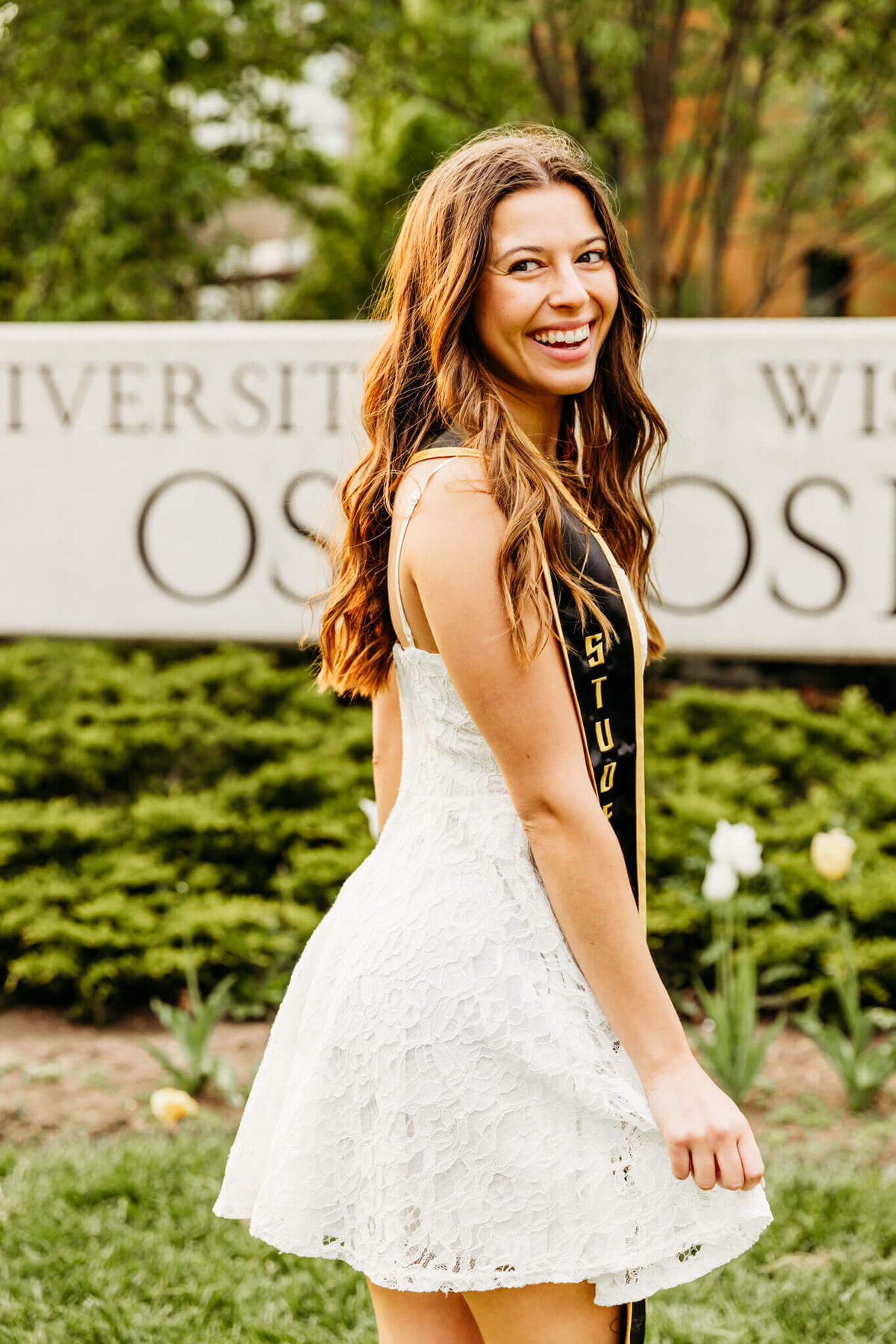 Stunning woman celebrating her college graduation while wearing a white lace dress and laughing on UW Oshkosh campus