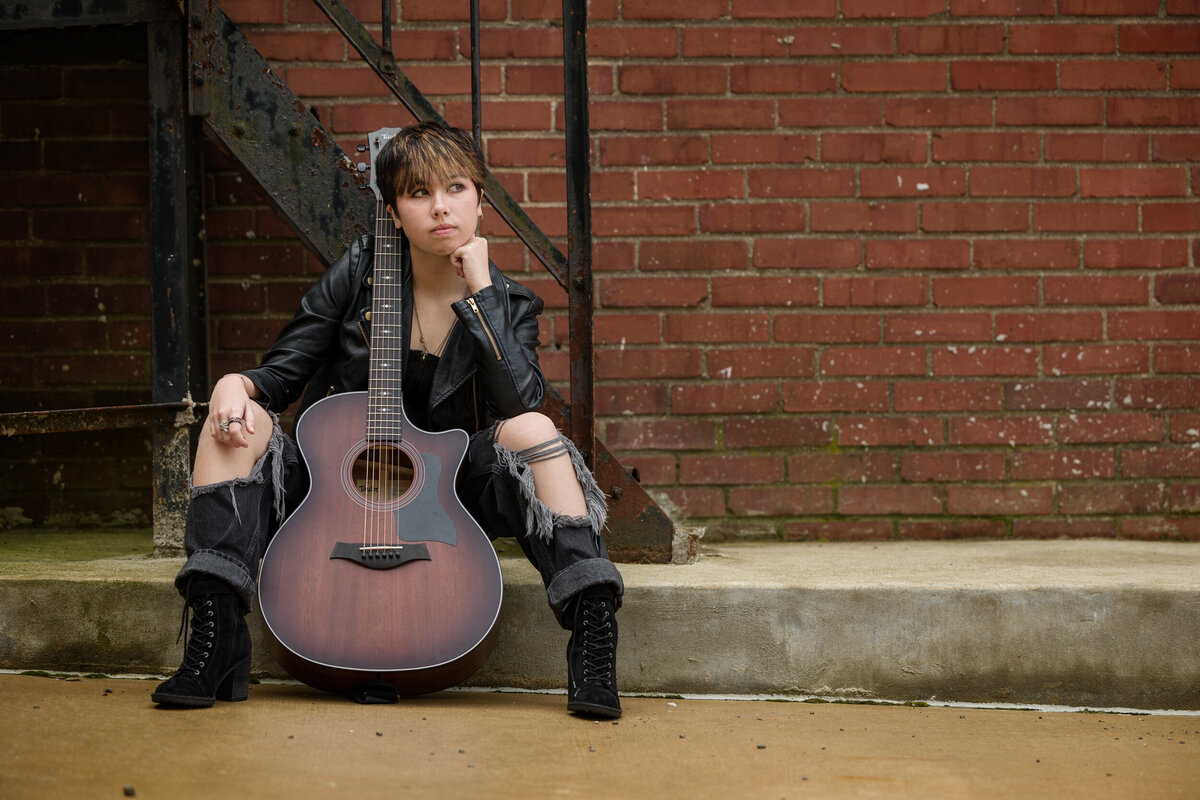 Branding photo of a young musician sitting with a guitar outside leaning against a brick wall