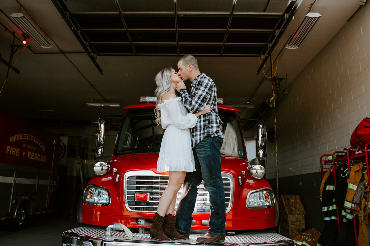 Engagement photoshoot at London, Ontario fire station. Man and Woman are standing on the front of the fire truck kissing. The woman has her hands around the man's middle and the man is lifting the woman's chin up for a kiss.