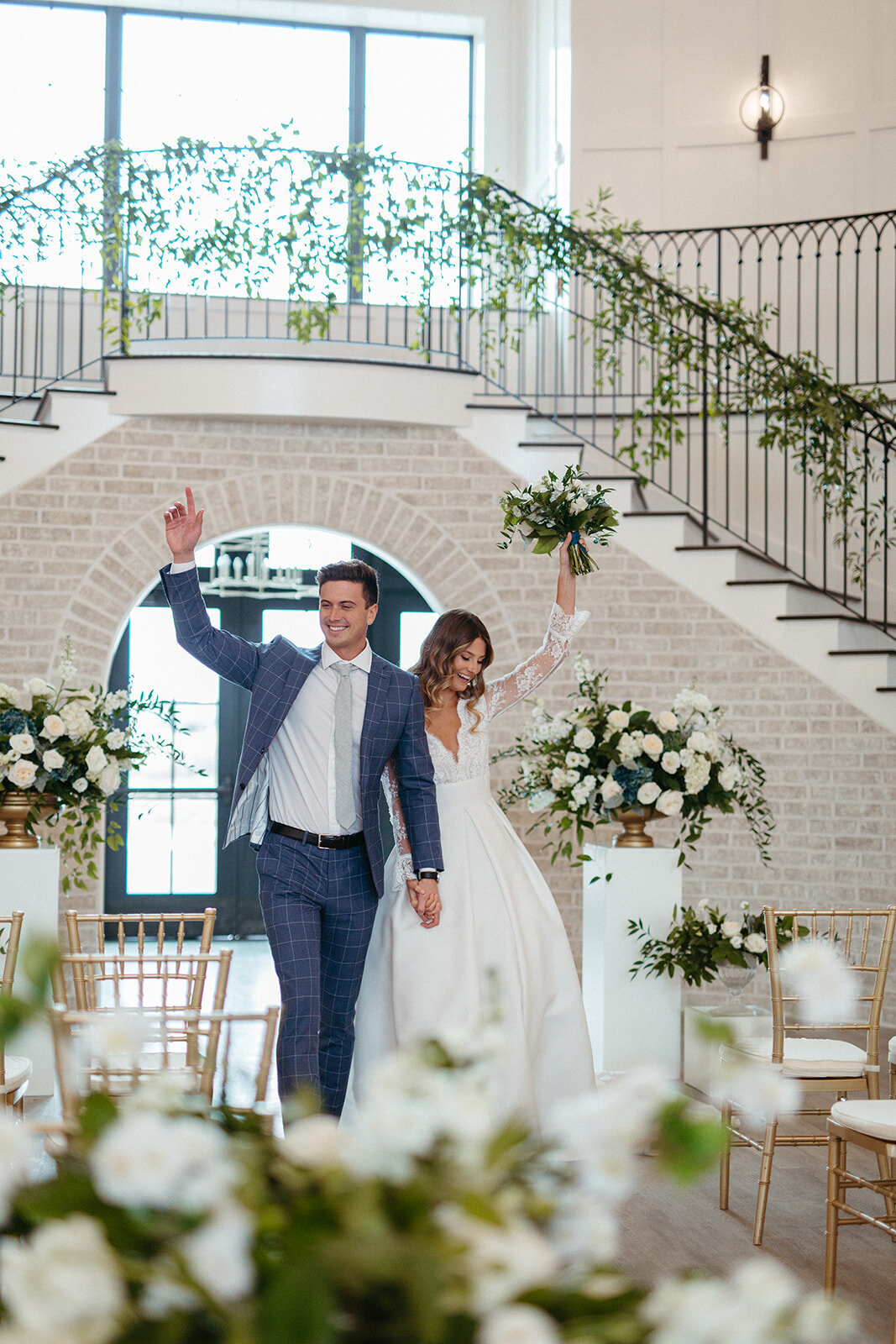 Bride and groom in a blue suit and white wedding gown wave walking down an aisle of gold chairs and white flowers.