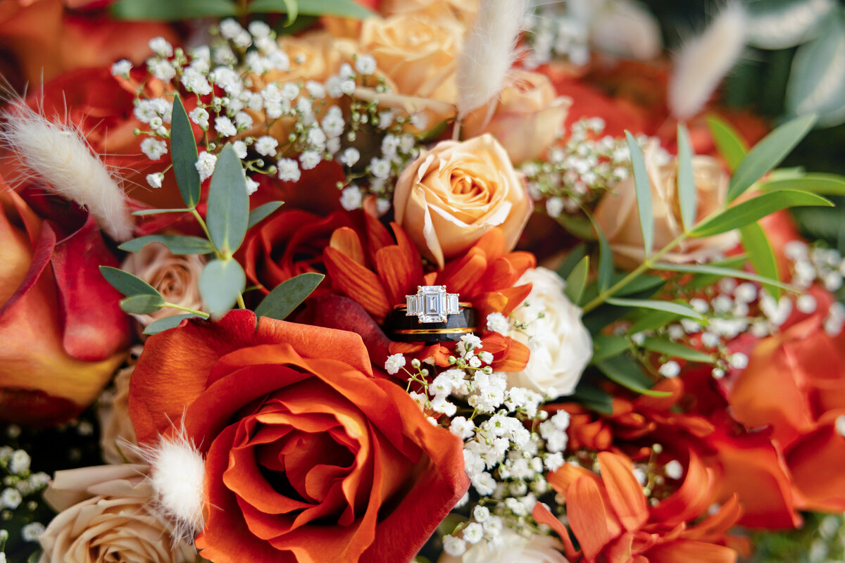  A close-up of a bridal bouquet with burnt orange and cream roses, featuring the couple’s wedding rings nestled in the petals.