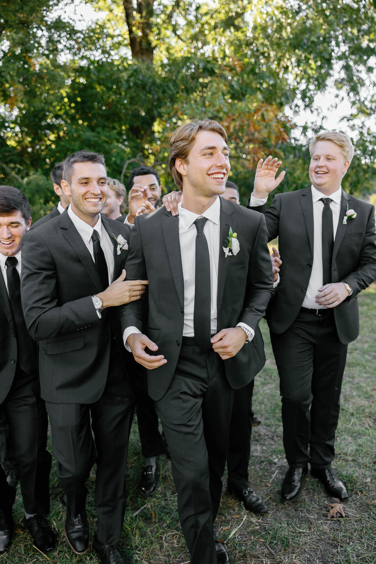 Groomsmen in black suits celebrating outdoors before the ceremony, each wearing coordinated white floral boutonnieres designed by the wedding florist.