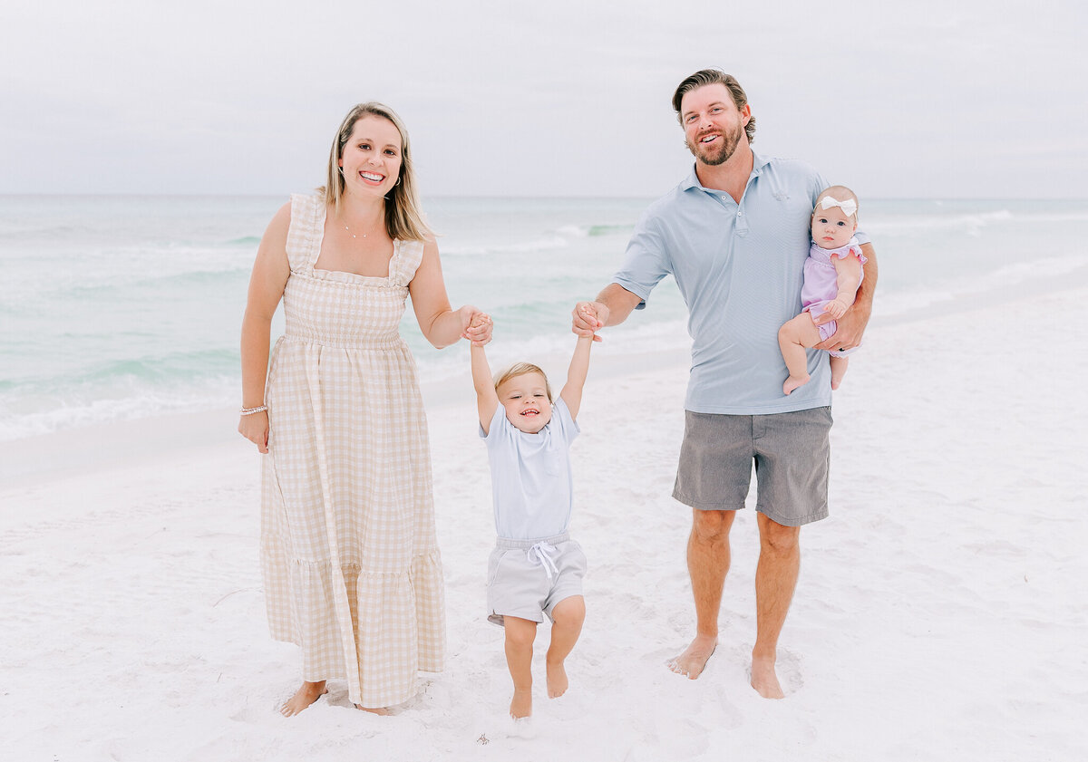 A joyful family of four enjoys a beach session on the white sands of Orange Beach, Alabama. The mother and father swing their young son between them while holding their baby daughter, capturing a fun and lighthearted moment by the ocean.