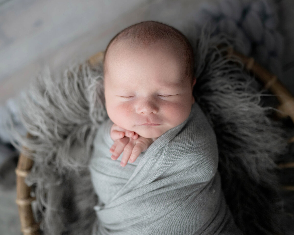 A baby sleeps in wicker bucket with fingers poking out top of grey swaddle