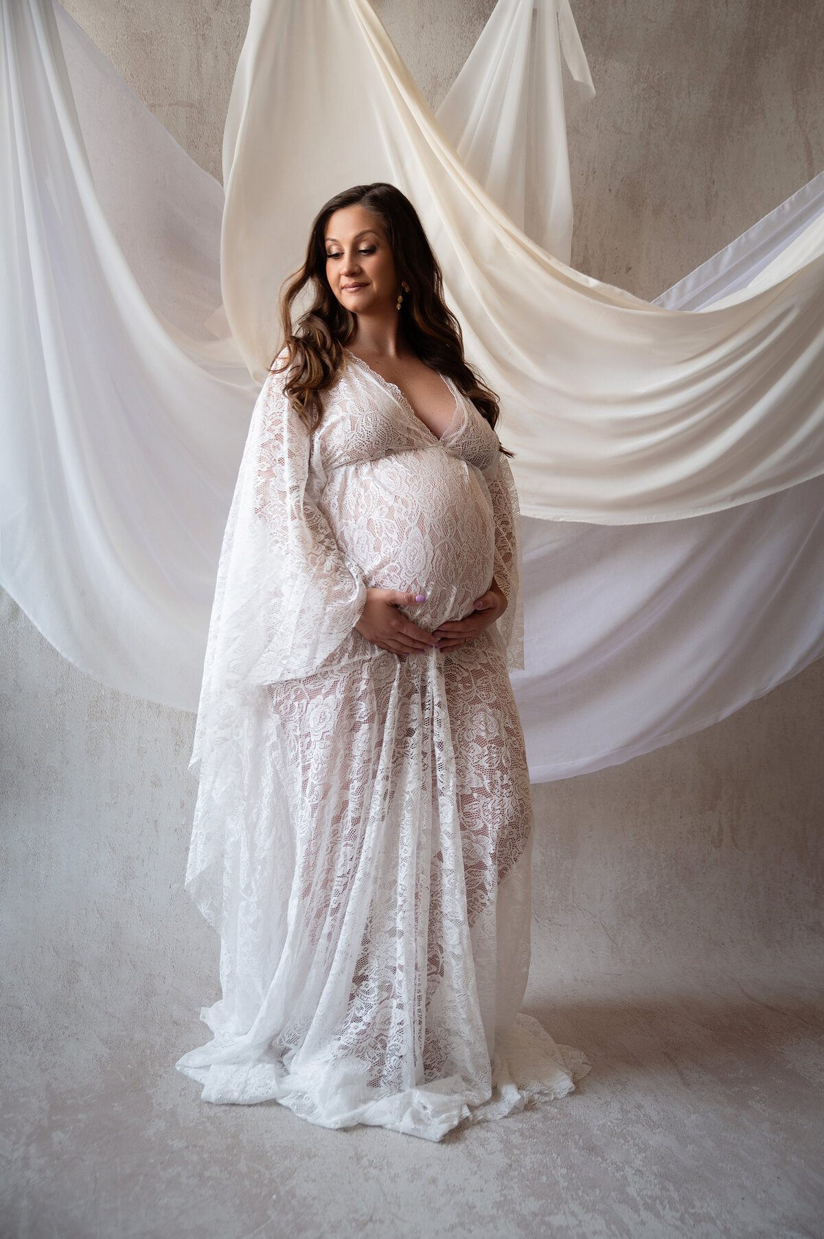 glowing pregnant brunette woman holding her baby bump wearing fitted boho cream maternity gown in front of cream and rustic backdrop and draped white fabrics