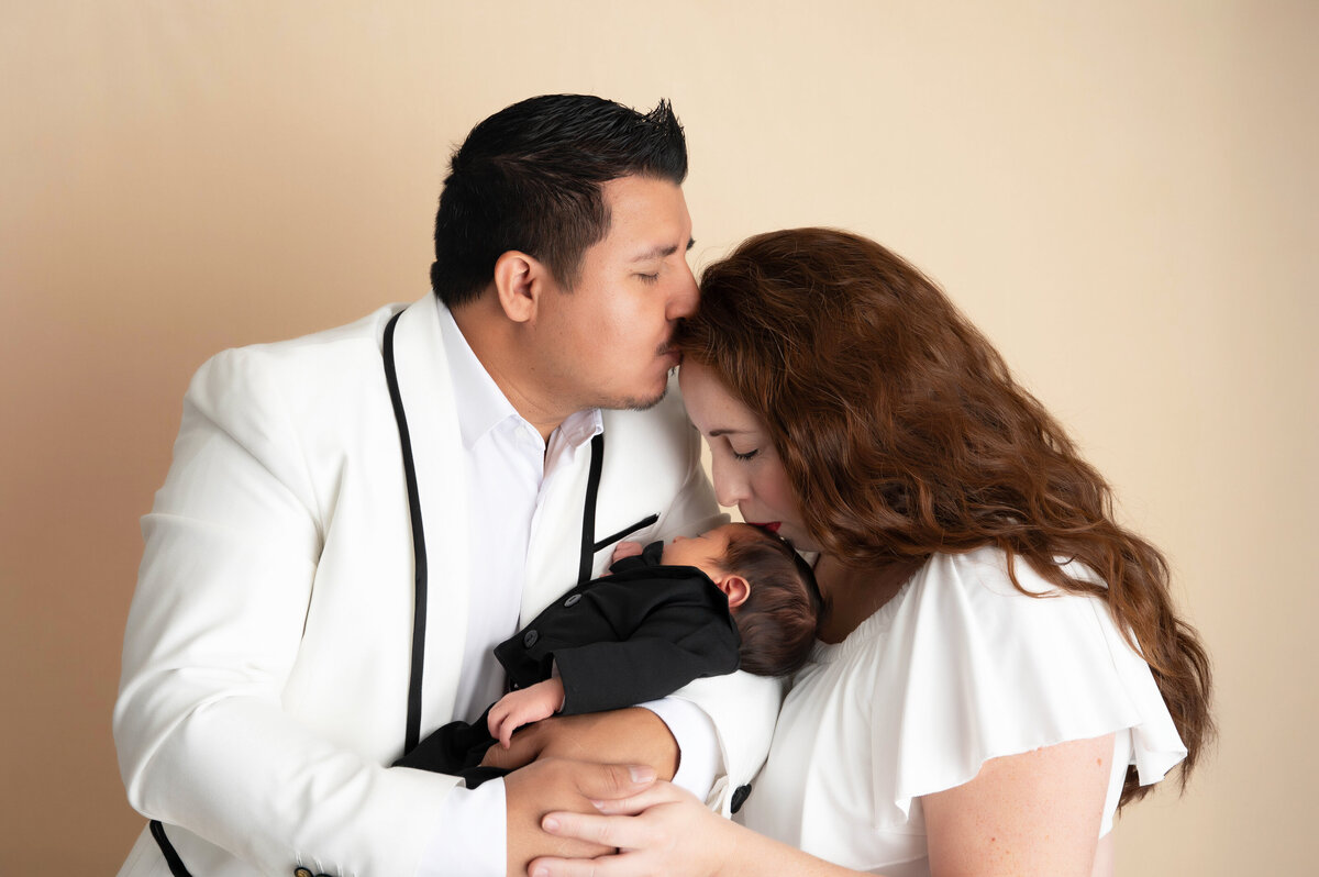 Close-up of a man holding a newborn baby, gently kissing a woman on the temple as she kisses the baby’s temple.