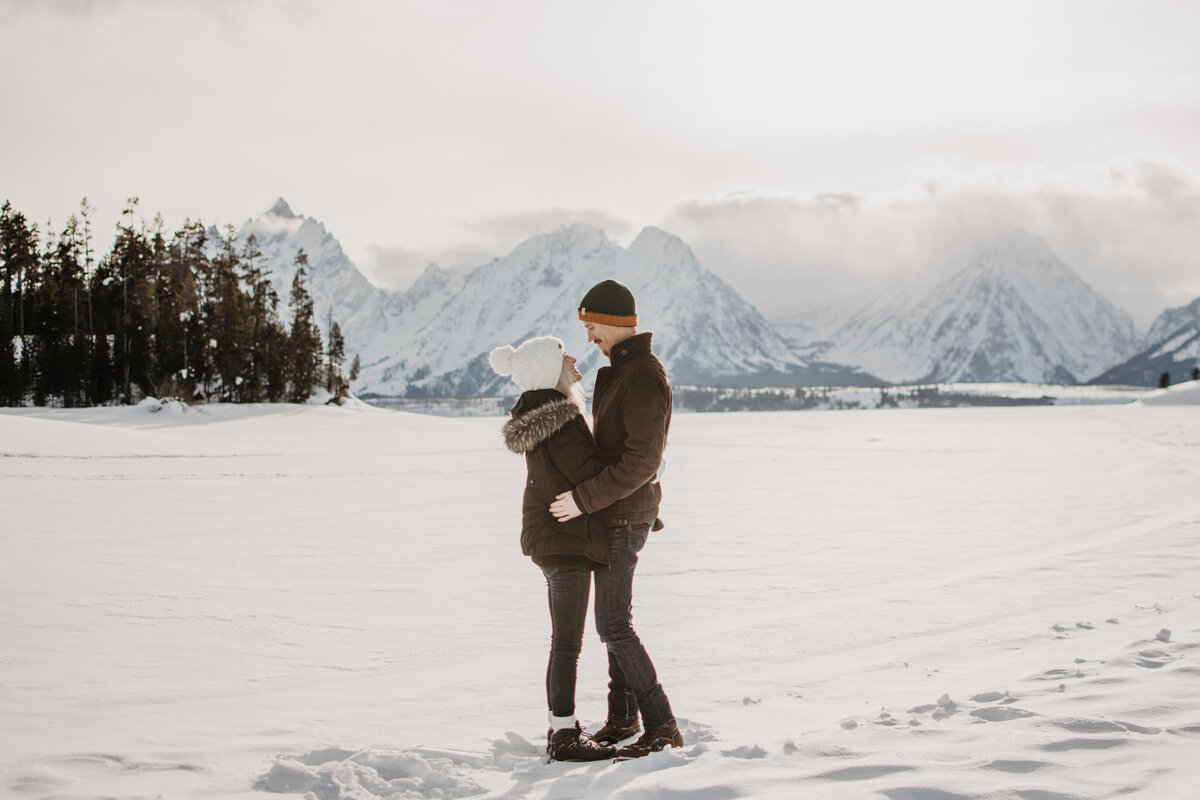 jackson hole photographers captures man and woman facing each other and holding each other by the hips as they standin gfront of the Grand Tetons for a winter engagement session in the snow