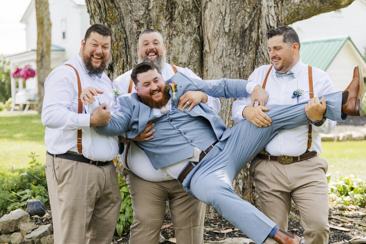 Groomsmen in suspenders laughing and lifting a groomsman off the ground on a lawn