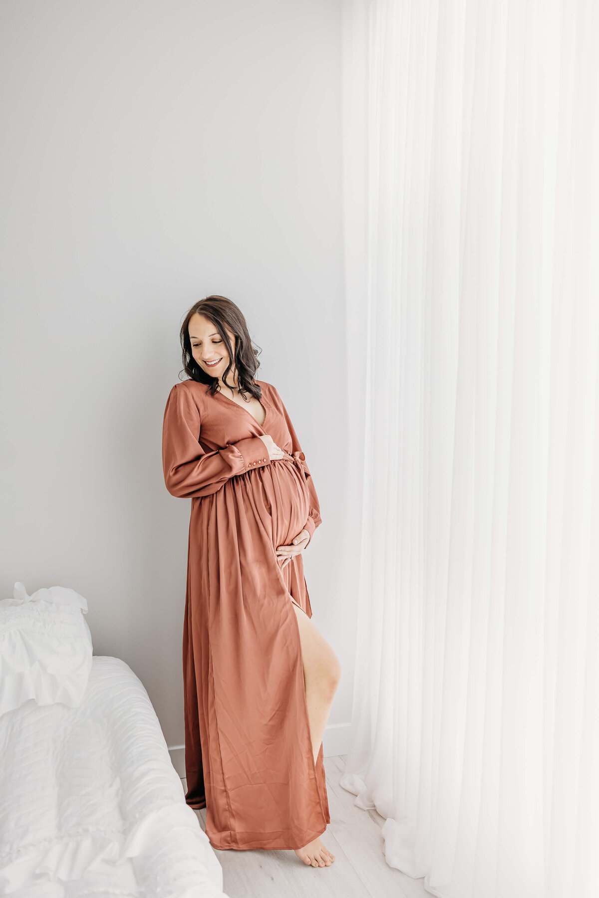 Happy mom with brown hair and dark pink gown standing near window for maternity pictures