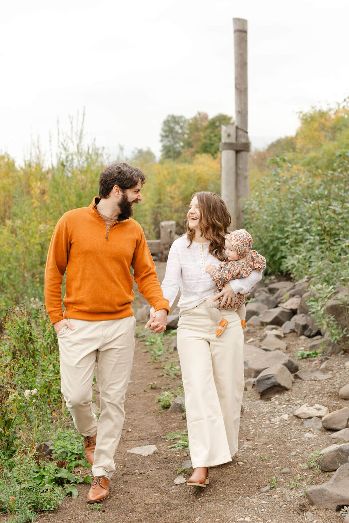 Mom and Dad are holding hands and walking together looking at each other out in nature. mom is holding baby on her hip. Dad is in a rust colored sweater and baby is wearing a floral romper with rust hues. Mom is in light, neutral tones. There is a lot of greenery to the sides and behind them.