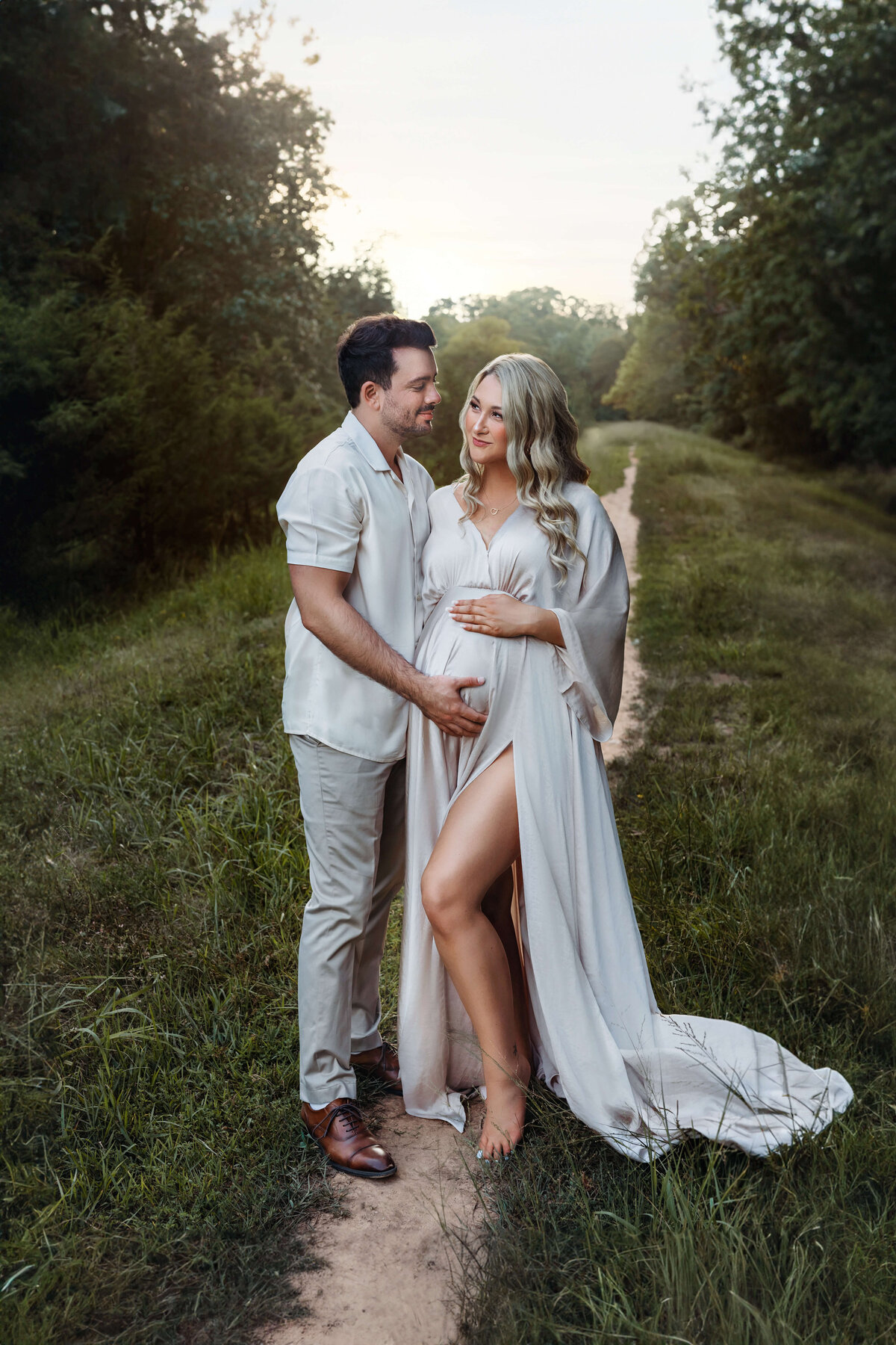 pregnant woman wearing a silky flowy sand colored maternity gown standing in a field holding her husband looking at eachother lovingly with the sun setting behind her in a  field at her northern va maternity photo session