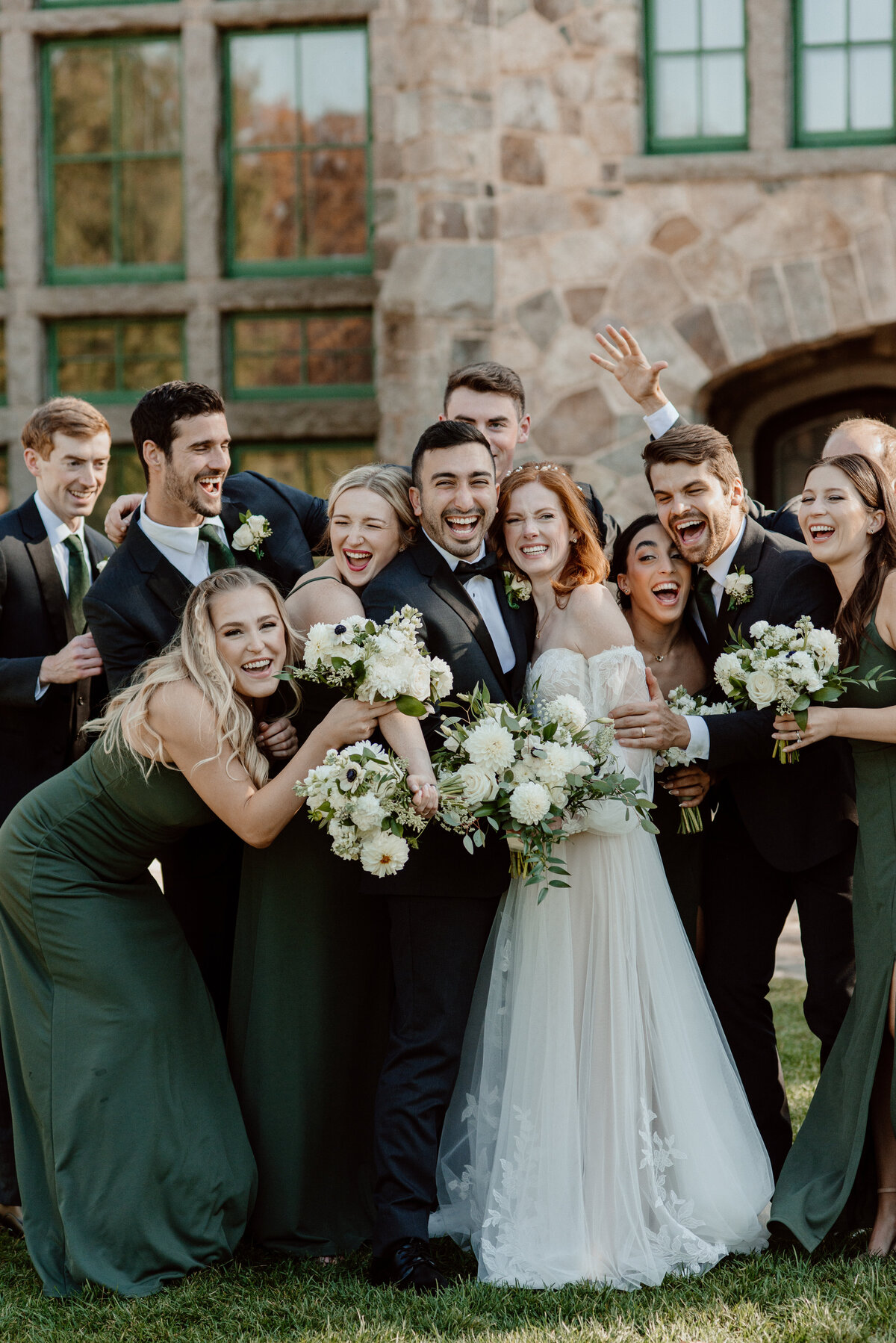 Joyful bride and groom surrounded by their enthusiastic bridal party at Borderland State Park in Easton, MA. The bride wears an off-the-shoulder gown, and the bridesmaids are in green dresses holding white bouquets. The groomsmen are in dark suits with green ties.