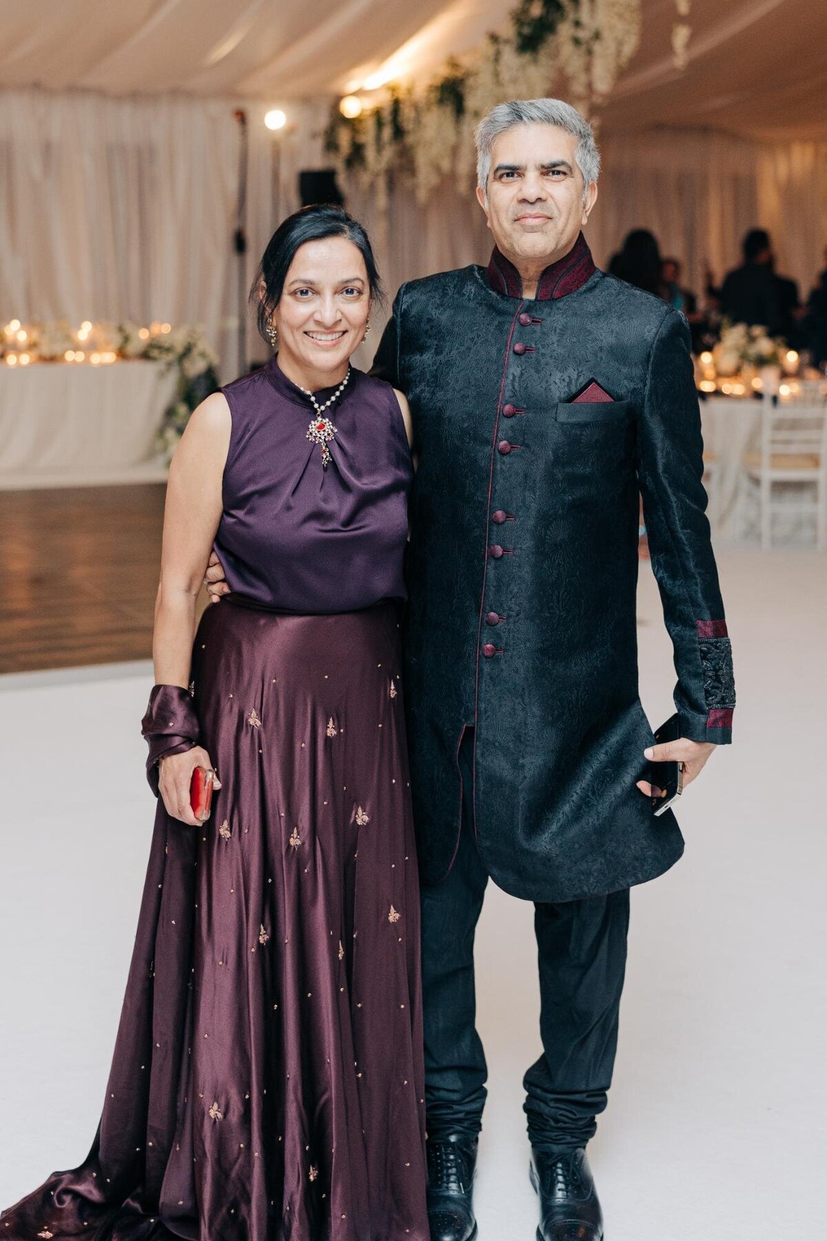 A couple in formal attire posing at an event; the woman in a purple gown and the man in a black traditional outfit with burgundy accents.