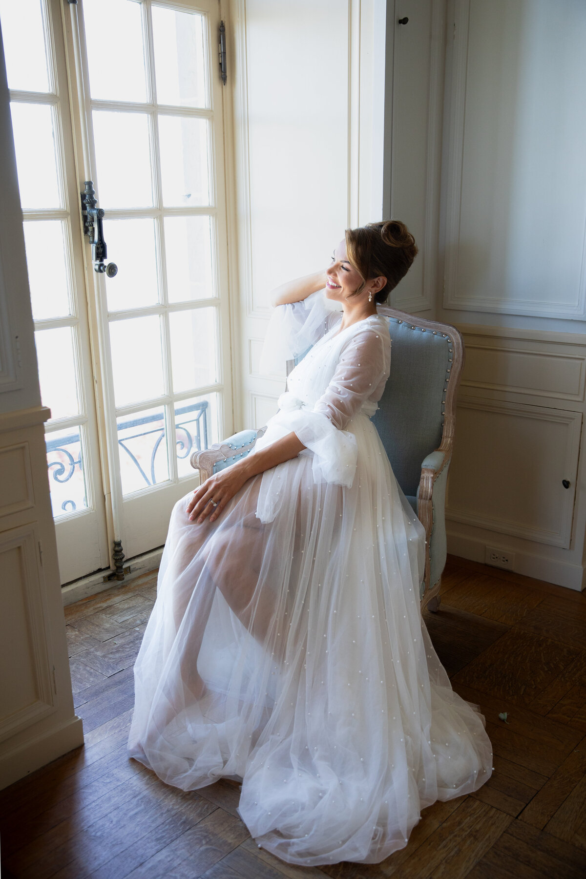 Rhode Island Wedding Photographers | Kelsey Sheehan Photography Timeless Rhode Island Weddings | A woman in a sheer white robe sits on a plush chair by a sunlit window. She smiles gently, conveying a serene and joyful ambiance.