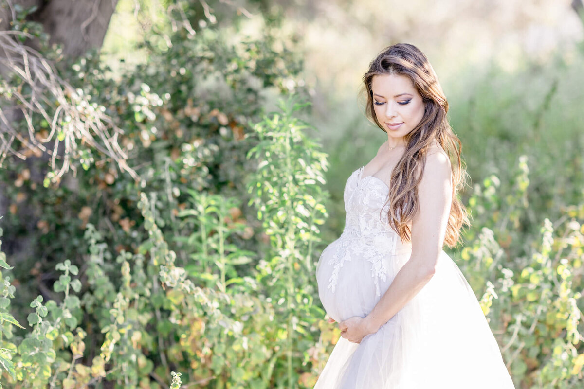 A long brown-haired pregnant woman in a long, white dress standing in a forest