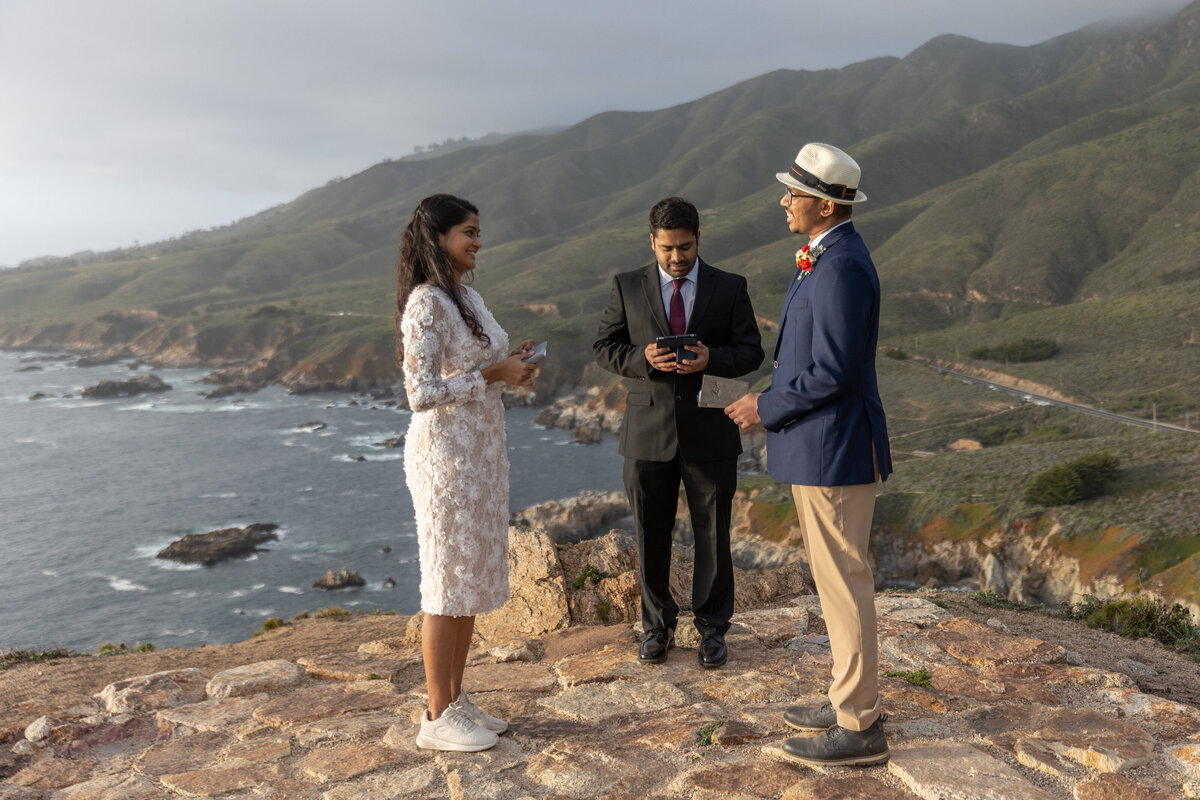 A bride and groom stand facing each other during their elopement ceremony in Big Sur with the ocean behind them.