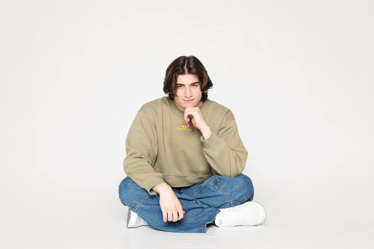 A person with medium-length dark hair sits cross-legged on the floor against a plain background. They are wearing a beige sweater, blue jeans, and white sneakers, resting their chin on one hand while looking at the camera.