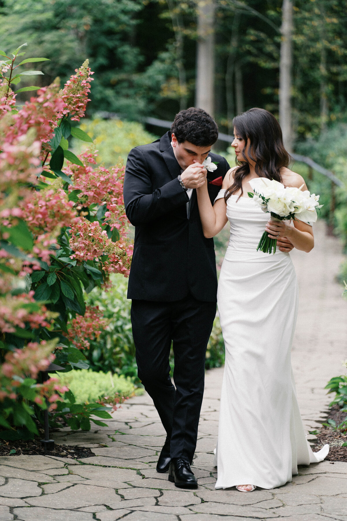 Groom kisses bride’s hand as they walk garden path at Sapphire Creek Winery wedding


