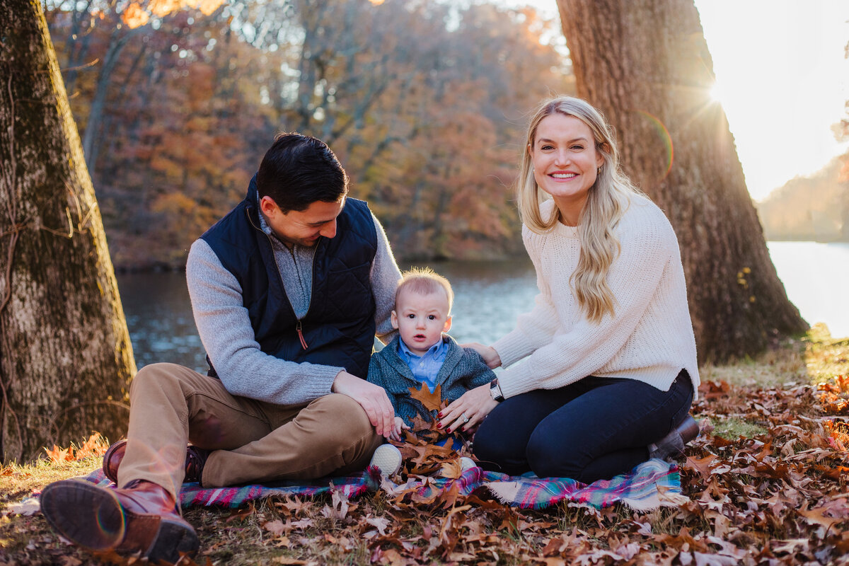 family of three sitting on a plaid blanket in the autumn leaves with a lake and setting sun behind them