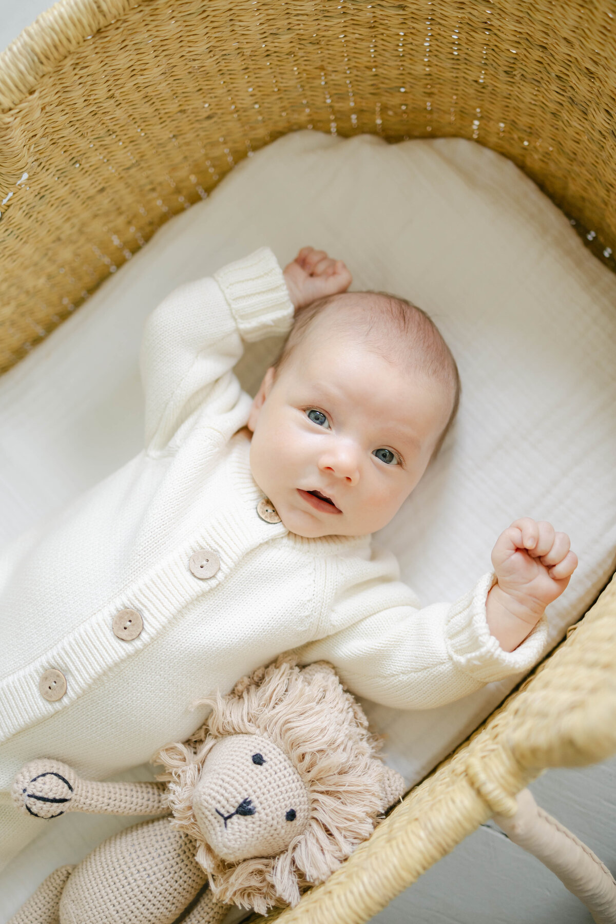 newborn baby looking at the camera wearing a beige knit romper with wooden buttons in a moses basket