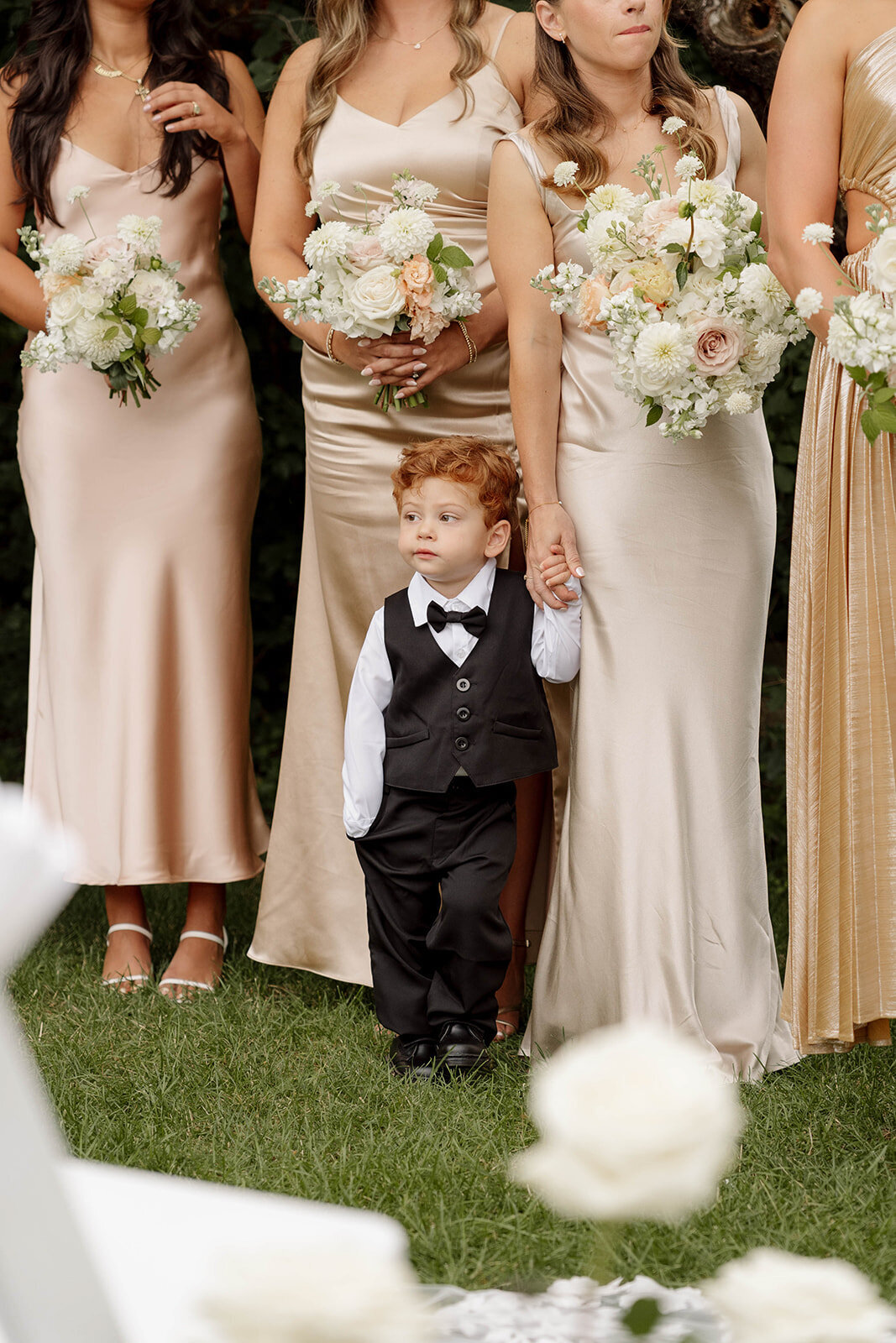 Bridesmaids in champagne dresses holding lush bouquets pose with an adorable ring bearer.