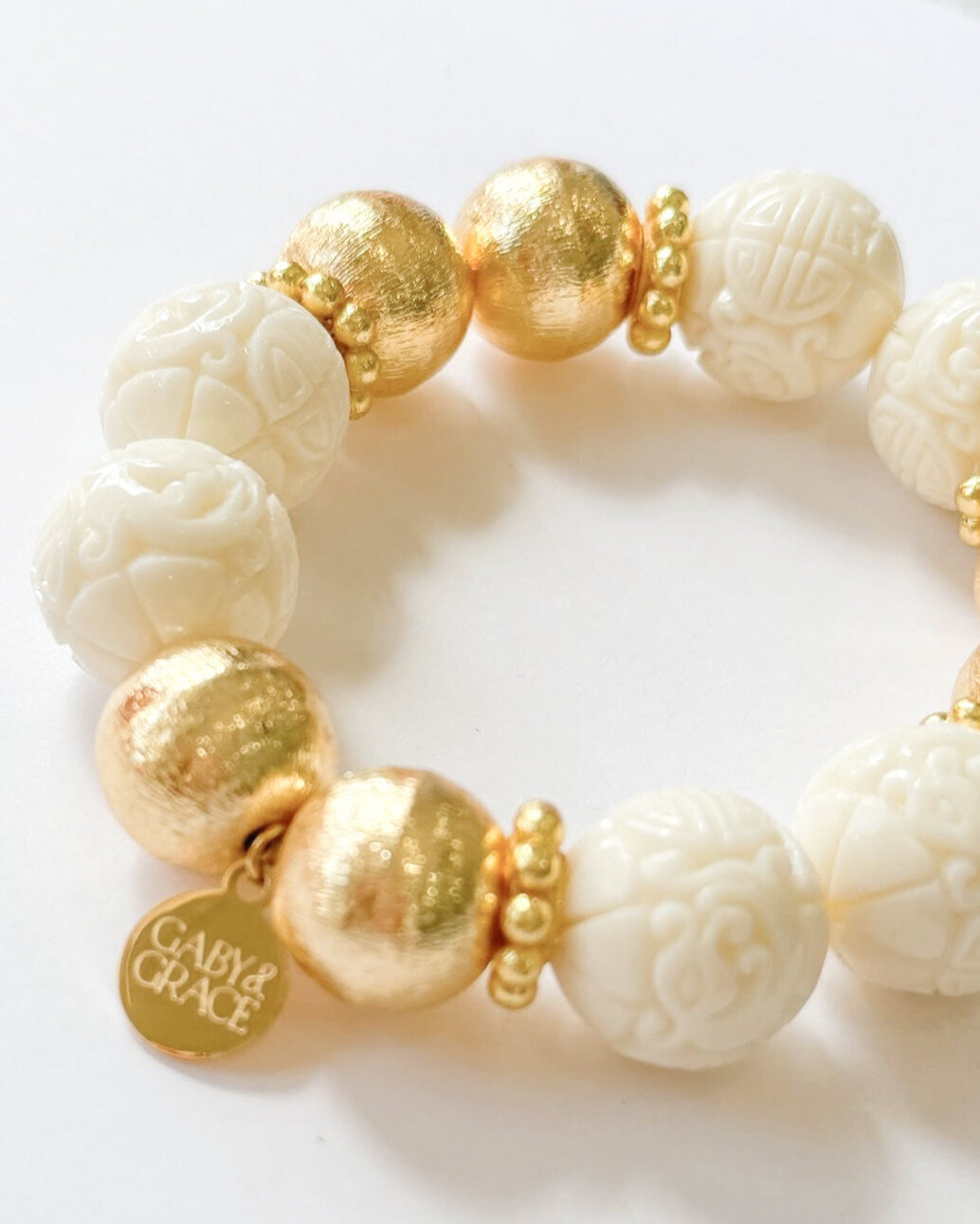 Close-up of an elegant Gaby & Grace bracelet featuring carved ivory-look beads and brushed gold rounds with a custom Gaby & Grace charm.