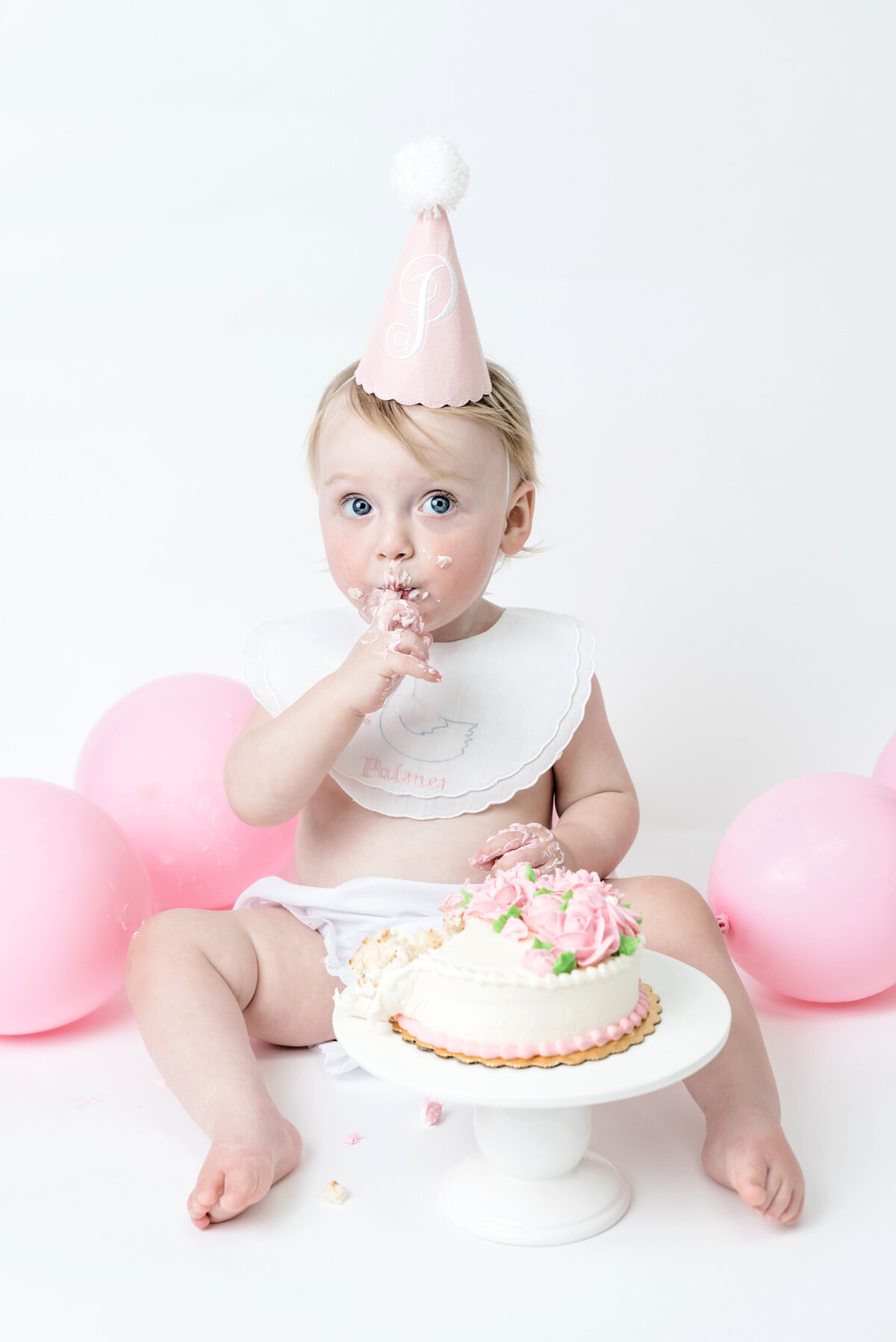 little girl wears pink hat and sits with cake and pink balloons during first birthday session at studio