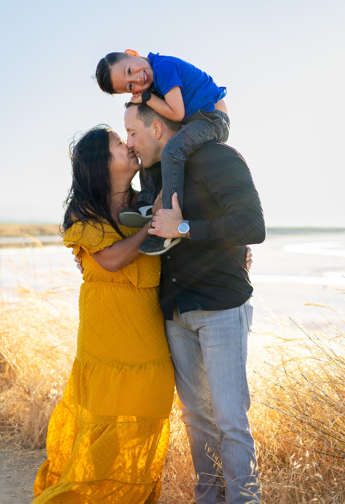 Young boy smiling from dad’s shoulders as parents share a kiss in tall golden grass – Bay Area Family Portfolio - Ellobelle Photography