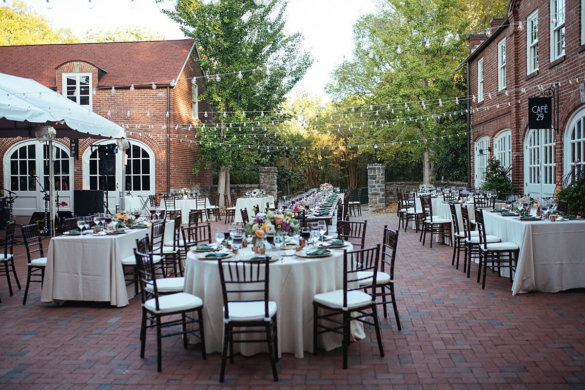Wedding reception tables set up at the exposed brick courtyard of the Frist Learning Center at Cheekwood Botanical Gardens. Tables are set with ivory table cloths and brown chiavari chairs while string lighting hangs above them.