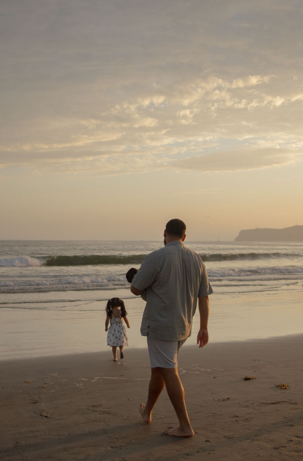 Father walking along the shoreline with his daughter, carrying his newborn baby, while watching the waves
