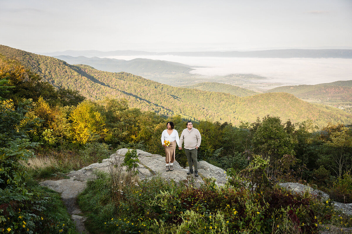 A couple stands on a rock with a mountain in the background. They are photographed in Shenandoah National Park by a Virginia elopement photographer.