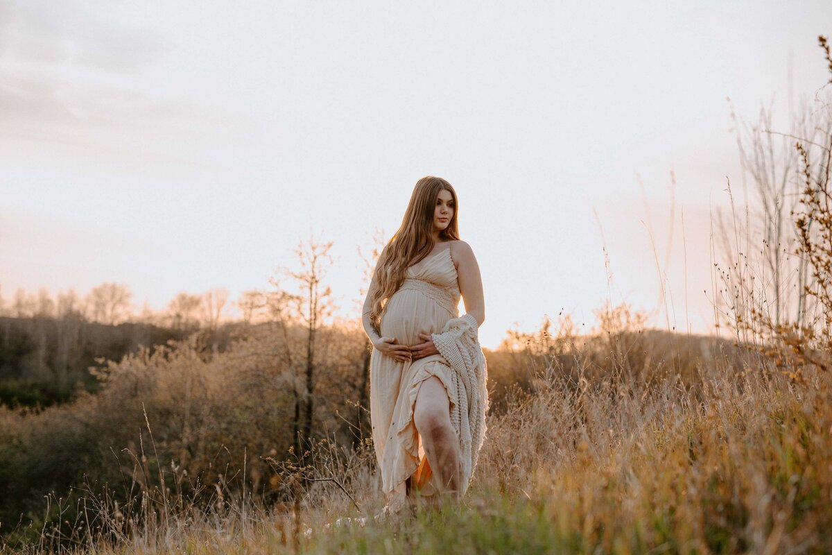 Expectant mom in Exeter, Ontario maternity photo shoot. She is walking in long grasses on rolling hills, hands touching her bump and looking off in the distance.