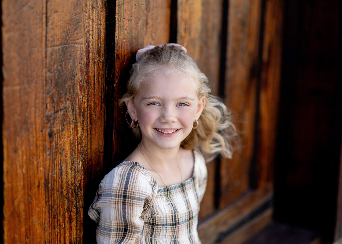 A child stands alone against a warm wooden wall, their face beaming.
