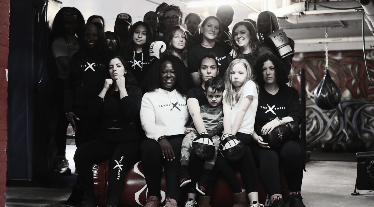 A diverse group of women and children gathered in a boxing gym, wearing Femme X Boxing branded clothing. They pose together in front of colorful wall art, some holding boxing gloves, with expressions ranging from determined to joyful. The image captures the inclusive community spirit of Femme X Boxing's programs that welcome participants of various ages and backgrounds