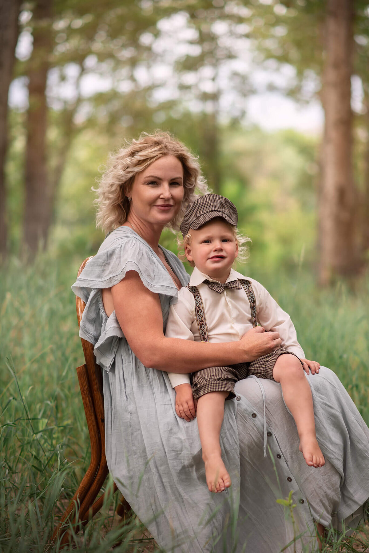 A mother wearing a flowing blue dress holds on her lap a toddler boy dressed in suspenders and pageboy cap. Taken in Kamloops by Brittany Danielle