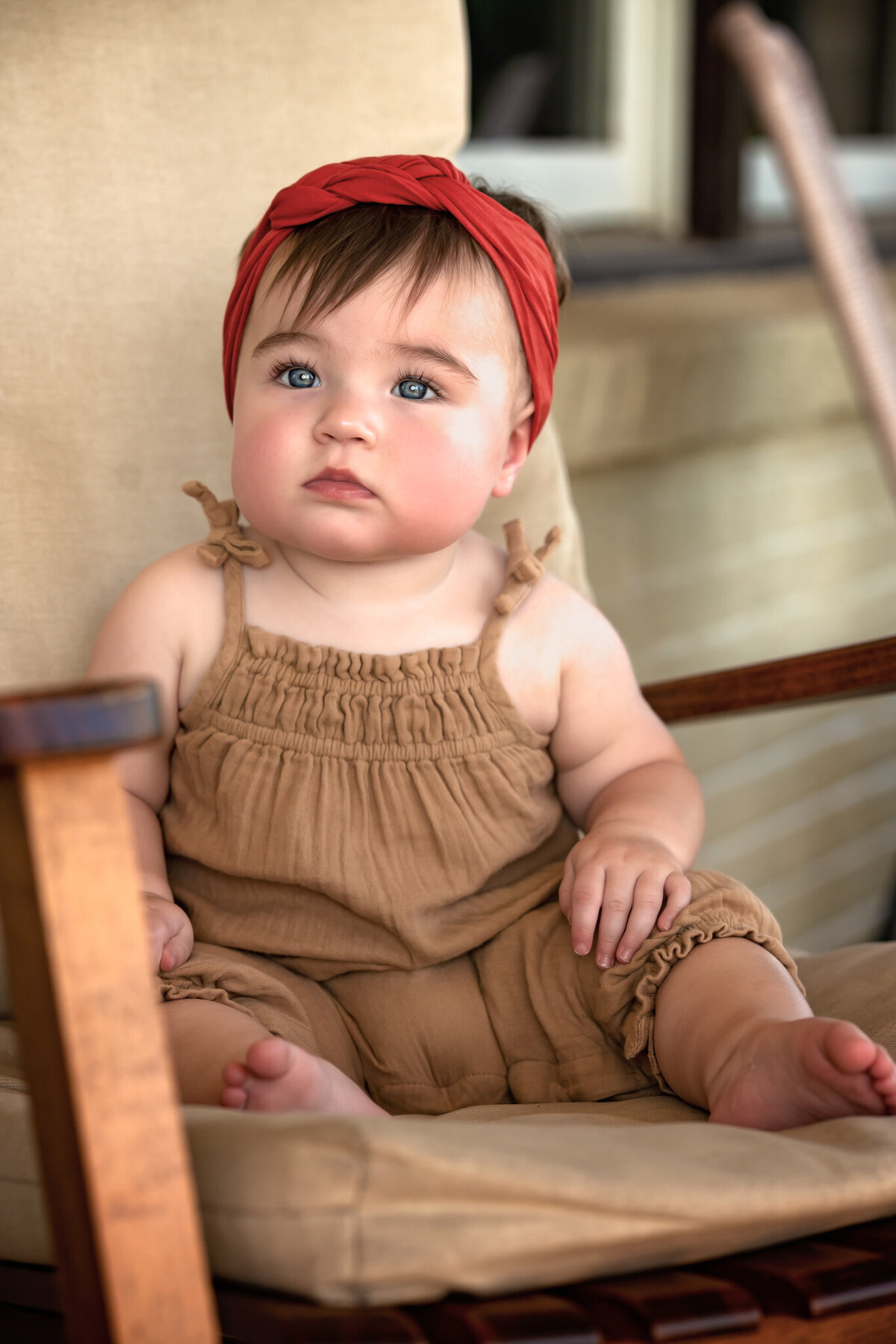 A baby girl is wearing a red headband and sitting on a wooden chair.